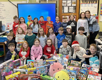 A group of students and a teacher gathered in a classroom, surrounded by a variety of toys and games. The classroom is decorated with educational posters and a whiteboard is visible in the background. Everyone is smiling at the camera.