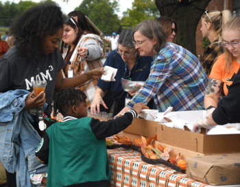 Person handing a snack to a child at a community event, with several people around a table filled with food.