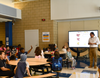 A presenter standing beside a digital screen displaying a map addresses students seated on the floor in a school cafeteria.