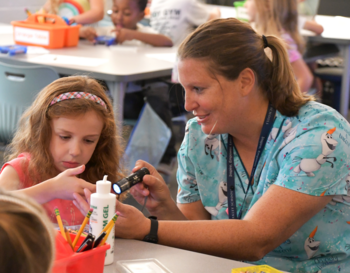 Nurse Rose and a young student are engaged in an activity at a classroom table, with the educator assisting the student with a germ spotting flashlight. Both individuals are focused on the task with school supplies visible around them.