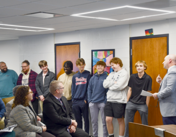 A group of individuals of mixed ages listens attentively to a speaker in a board office adorned with artwork. Some participants are smiling.