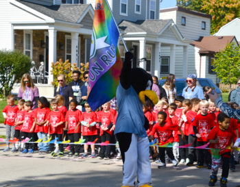 Person in a penguin costume waving a rainbow flag at a starting line where children wearing red t-shirts are gathered for a race, with onlookers in the background.