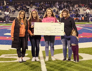 Five individuals presenting a large ceremonial check for $2,500 to Gilda's Club, standing on a football field with a midfield logo visible underfoot.