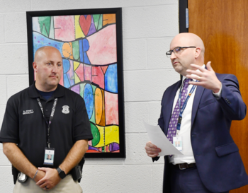 Two individuals in a professional setting, with one holding papers and speaking while the other listens attentively. They are standing next to a colorful artwork.