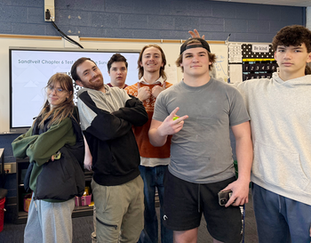 Group of five students and their teacher smiling in a classroom, with a whiteboard visible in the background.