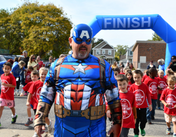 A person dressed as Captain America leading a group of children in red shirts through a 'FINISH' archway at a school event.