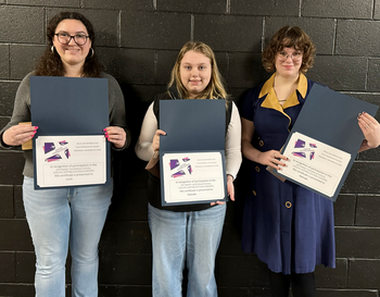 Three individuals holding certificates against a black brick wall, smiling proudly. Each certificate features a purple logo.