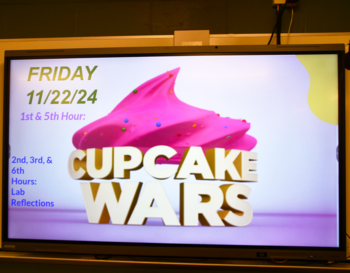 A digital display screen showing an announcement for 'Cupcake Wars' on Friday 11/22/24, listing schedule details beneath a large image of a pink, sprinkle-topped cupcake.