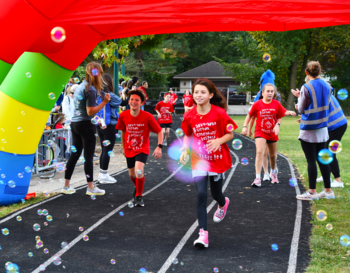 Participants in red shirts running through a large inflatable rainbow arch at a charity event, surrounded by floating bubbles and spectators cheering.