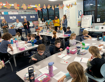 A classroom scene with multiple children seated at tables, engaging in various activities. Adults are standing, supervising and assisting. The room is decorated with educational posters and colorful craft papers.