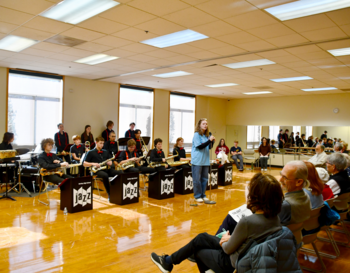 An indoor concert presentation by a school band featuring various musicians playing instruments like drums and saxophones, with an audience of all ages watching and listening attentively.