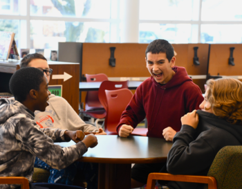 Four students sit around a table in a library, engaged in an animated conversation. One student is laughing joyfully. Shelves with books and a large window can be seen in the background.