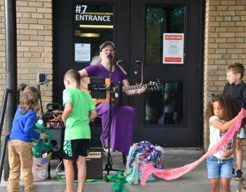 A musician playing a guitar and singing into a microphone at a school entrance labeled 