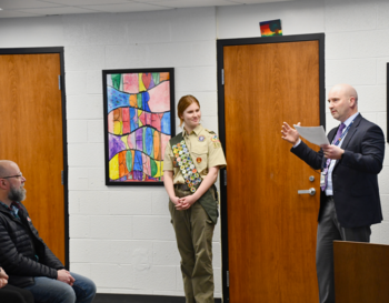 A person in a Scout uniform stands beside a door, listening to another person who is speaking and holding a paper. A seated individual observes the interaction in a room decorated with colorful wall art.