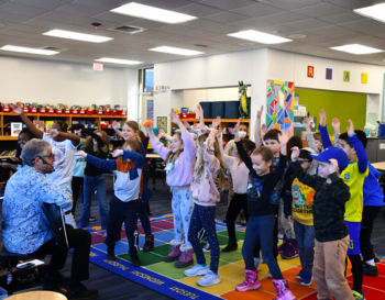 Adult leading a group of children in a stretching exercise inside a colorful classroom.