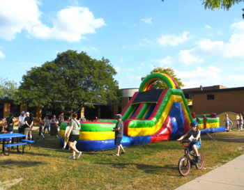 People enjoy a sunny day outdoors at a community event featuring a large, colorful inflatable slide. Some attendees are walking and biking along a path, with picnic tables nearby under clear blue skies.