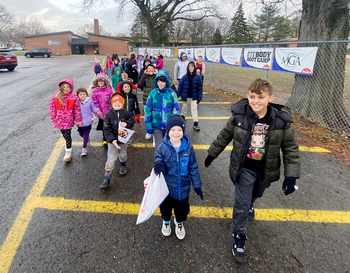 Group of children walking in a line on a paved path, smiling and dressed in winter clothing, with banners in the background.