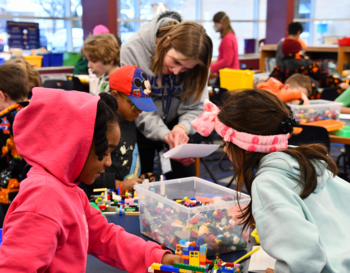 Children and an adult engage in building projects with LEGO bricks at a colorful activity table in a busy classroom setting.