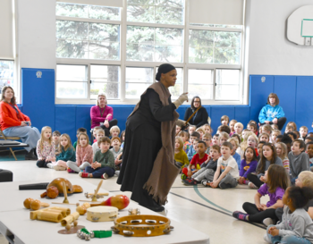 An individual dressed as someone traveling on the underground railroad demonstrates musical instruments to a group of attentive students seated on the floor of a school gymnasium.