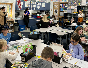 An elementary classroom scene with students seated at desks working on assignments. Three adults, possibly teachers, are present, instructing and assisting the students. The room is well-organized with educational posters and resources.