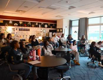 Students sitting in a classroom at desks, attentively watching a presentation. The room is decorated with educational posters