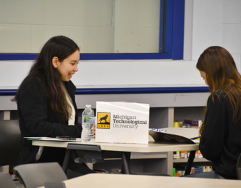 Two students seated at a table, with one smiling and looking at the other, in a room with a Michigan Technological University logo visible on a box.