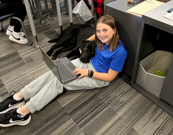 A person sitting on the floor of a classroom working on a laptop, with a black dog lying next to them. The classroom has desks with papers and books, and other students are visible in the background.