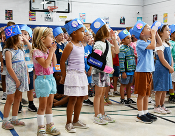 Children in a gymnasium wearing paper hats and holding signs, actively participating in an International Peace Day event.