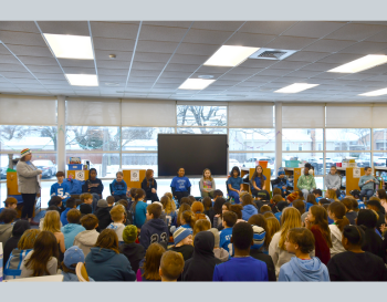 A group of students seated in a media center, attentively watching a presentation by several individuals standing in front of a large monitor.