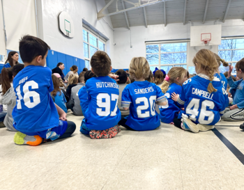 A group of children sitting on the floor in a gymnasium, three wearing blue lions jerseys with the names 