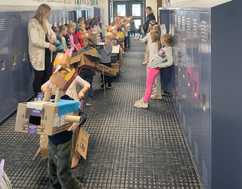 Children wearing cardboard costumes during a school activity in a hallway, with teachers present.