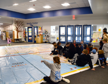 A group of students seated around a large floor map in a school's media center, engaged in a geographical learning activity.