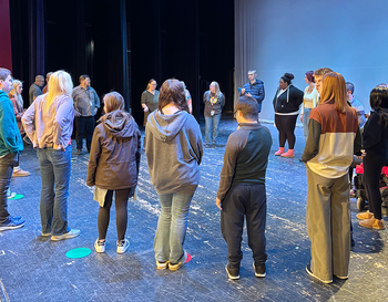 A group of people standing in a circle on a theater stage, participating in a workshop.