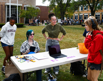 Four students interacting at a high school club recruitment table outdoors, with one seated and three standing. The table is labeled with a banner and features a laptop and promotional materials.