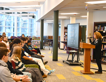 Person speaking at a podium in a library with an audience of students listening and a presentation being displayed on a screen.