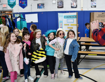 A group of children dressed in various costumes posing for a photo in a school gymnasium, with an adult taking their picture.