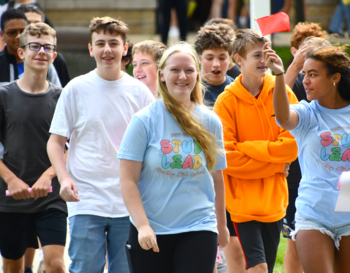 Group of students participating in an outdoor activity, some wearing 'STUD LEAD' t-shirts, smiling and walking on a grassy area.