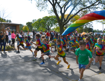 Children in colorful outfits participate in a fun run under a rainbow-colored arch on a sunny day.