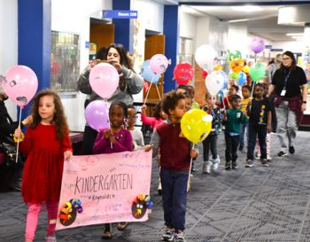 Group of children holding balloons and a banner that reads 'Kindergarten' as they walk through a school hallway, accompanied by adults.