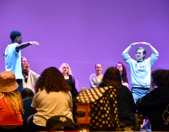 Two individuals performing on stage in front of an audience, with one person extending an arm towards the other who is forming an arch with their arms above their head. The background is illuminated in purple, and several spectators are visible enjoying the performance.