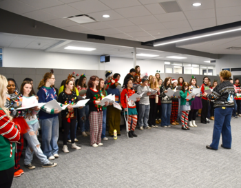 A group of people wearing festive holiday sweaters and accessories singing carols from sheets of paper.