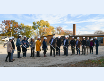 People in hardhats holding shovels in a construction zone tossing dirt during a groundbreaking ceremony