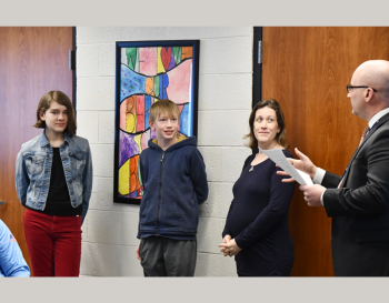 Three individuals listening to another person speaking, standing in a room with a colorful abstract painting on the wall.