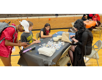 People in various costumes are crafting at a table in a gymnasium decorated for Halloween.