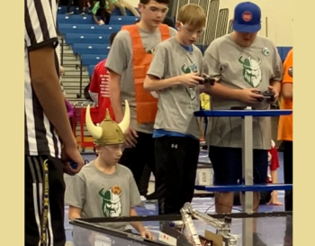 Three participants at a robotics competition, one wearing a horned hat, operate their controllers focused on a robotic device in the foreground.