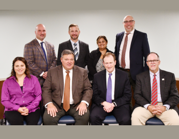 Group of eight professionals posing for a photo in a meeting room. Four individuals are standing in the back row, and four are seated in the front row. All are dressed in business attire.