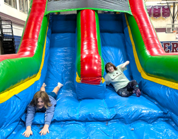 Two children are joyfully sliding down a colorful inflatable slide.