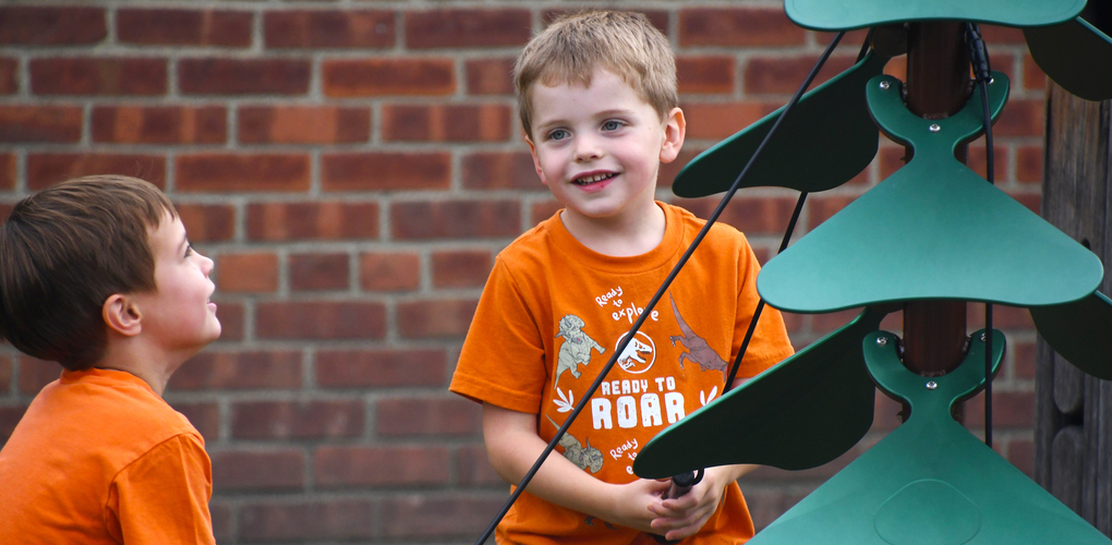 Two children playing outdoors with a colorful wind spinner, one holding the spinner pole and smiling, with a brick wall in the background.