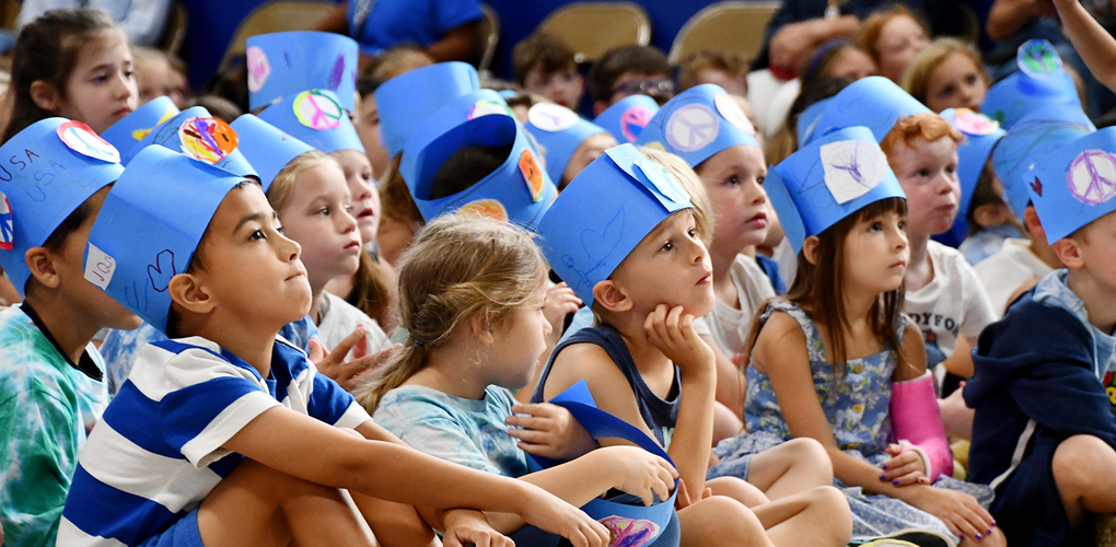 Group of children wearing blue paper hats sitting on the floor at a school event, listening attentively.