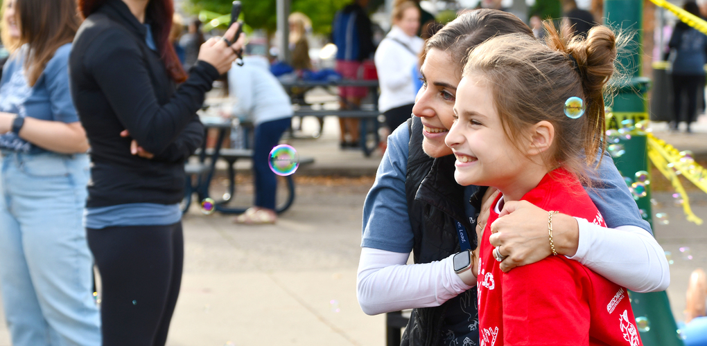 A joyful scene at an outdoor event with two people in focus at the center: an adult and a child warmly embracing and smiling, surrounded by floating bubbles. Both are casually dressed, highlighting a cheerful, communal atmosphere.